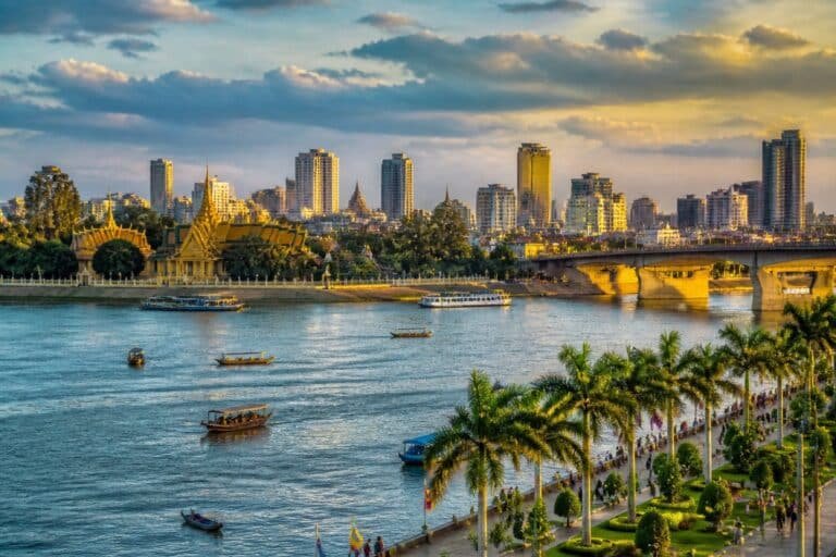 Vista panorâmica de Phnom Penh com o rio, barcos, palmeiras, o Palácio Real e edifícios da cidade ao fundo.