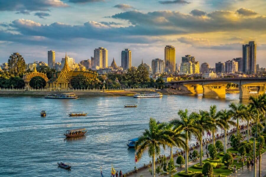Vista panorâmica de Phnom Penh com o rio, barcos, palmeiras, o Palácio Real e edifícios da cidade ao fundo.