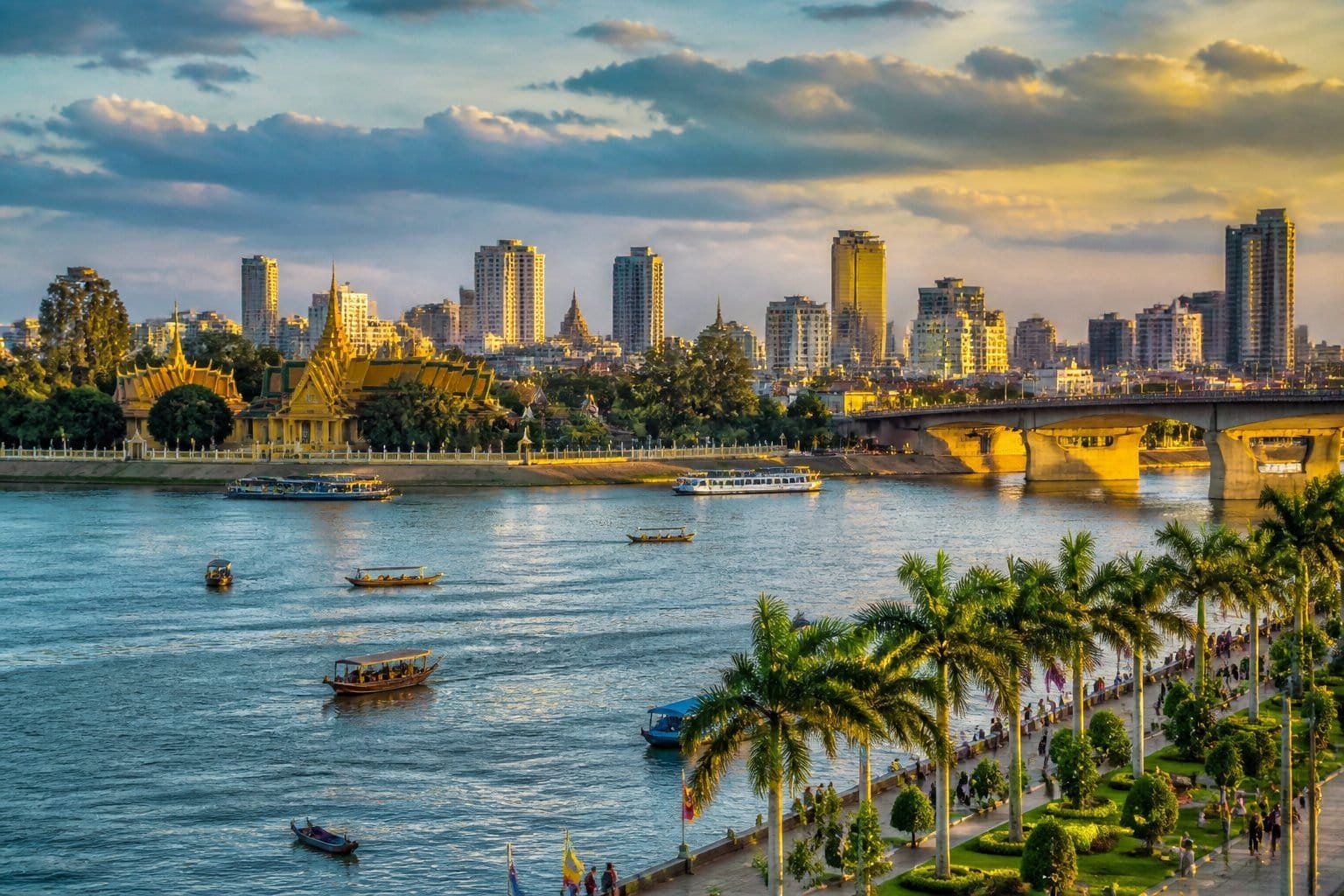 Vista panorâmica de Phnom Penh com o rio, barcos, palmeiras, o Palácio Real e edifícios da cidade ao fundo.
