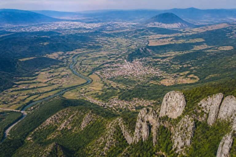 Paisagem montanhosa e rural dos Bálcãs, com vales, rios e cidades ao longe.