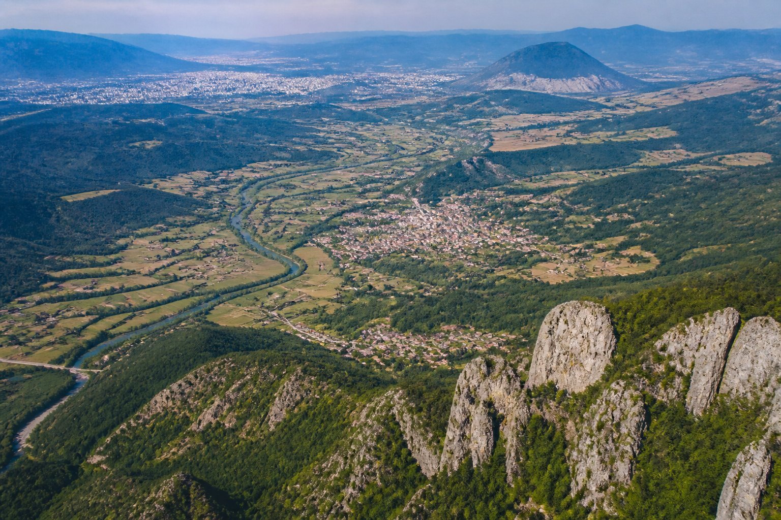 Paisagem montanhosa e rural dos Bálcãs, com vales, rios e cidades ao longe.
