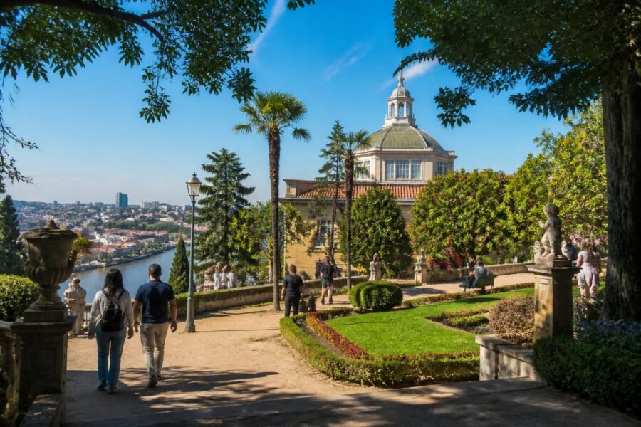 Visitantes passeiam pelos jardins do Palácio de Cristal, no Porto, cercados por árvores, flores e uma vista panorâmica.